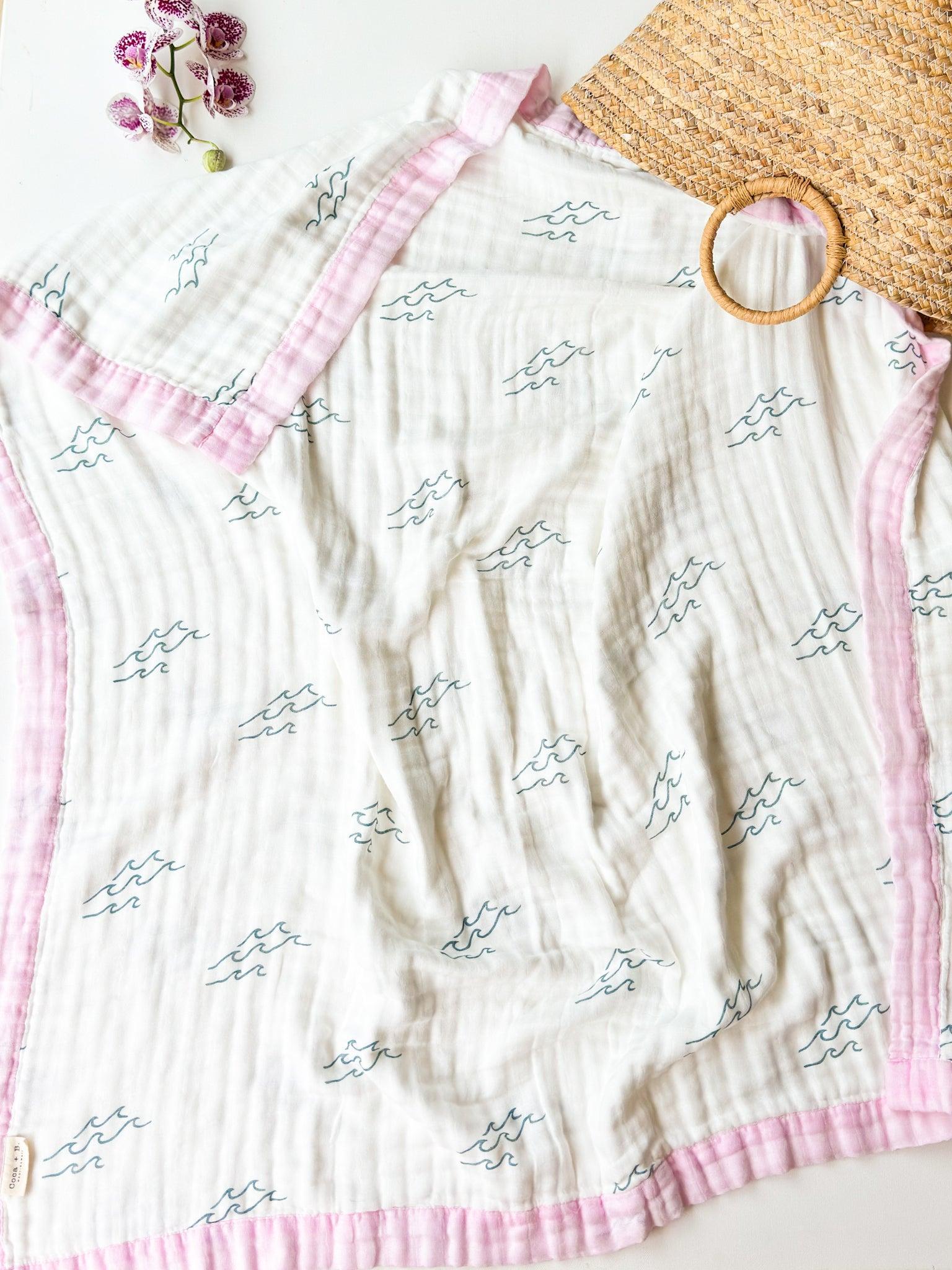 Baby blanket with pink trim and wave pattern on a white surface with a straw basket and flowers.